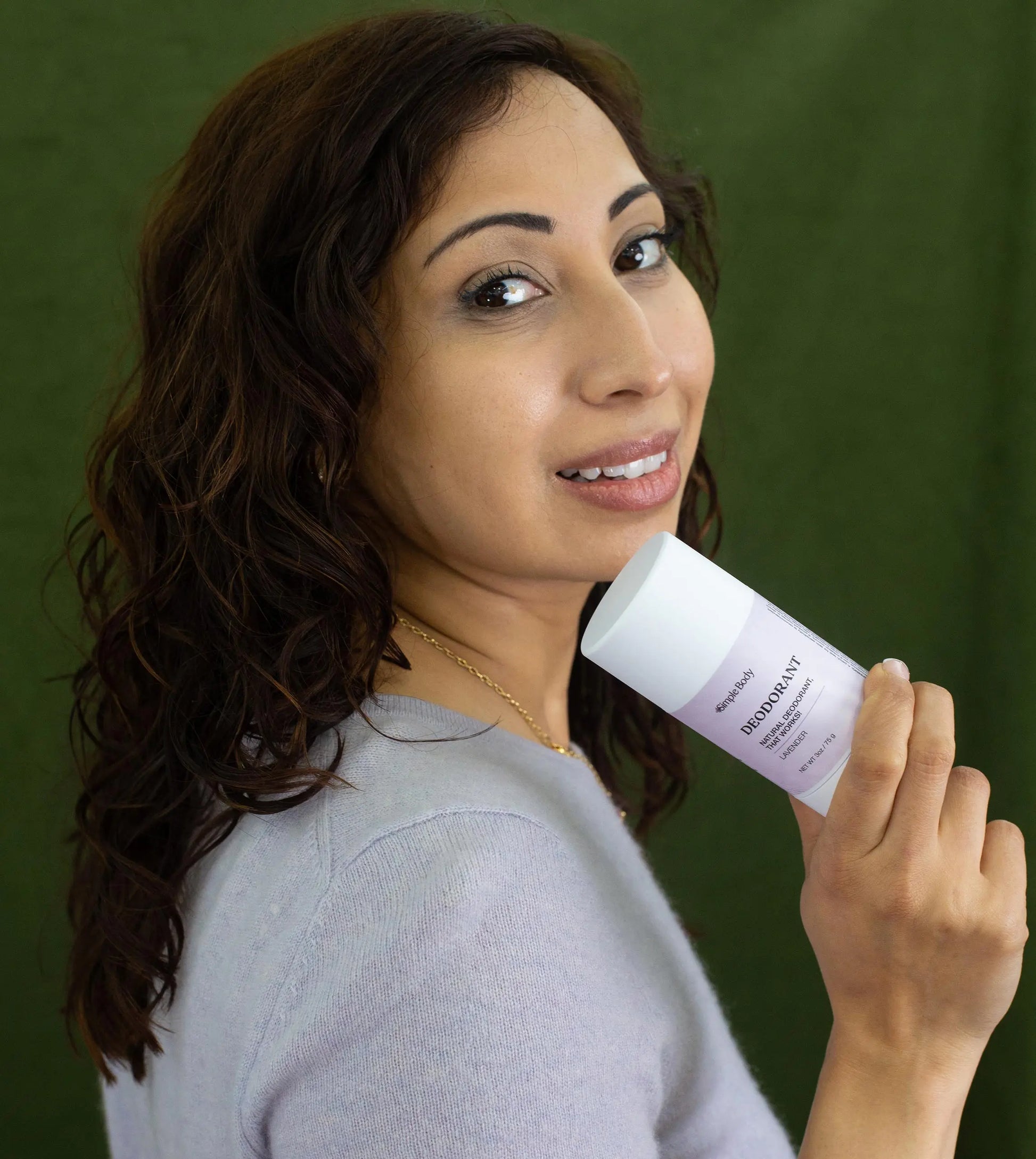 Woman holding a jar of Simple Body All Natural Lavender Deodorant looking over her shoulder with a purple sweater and a dark green background.