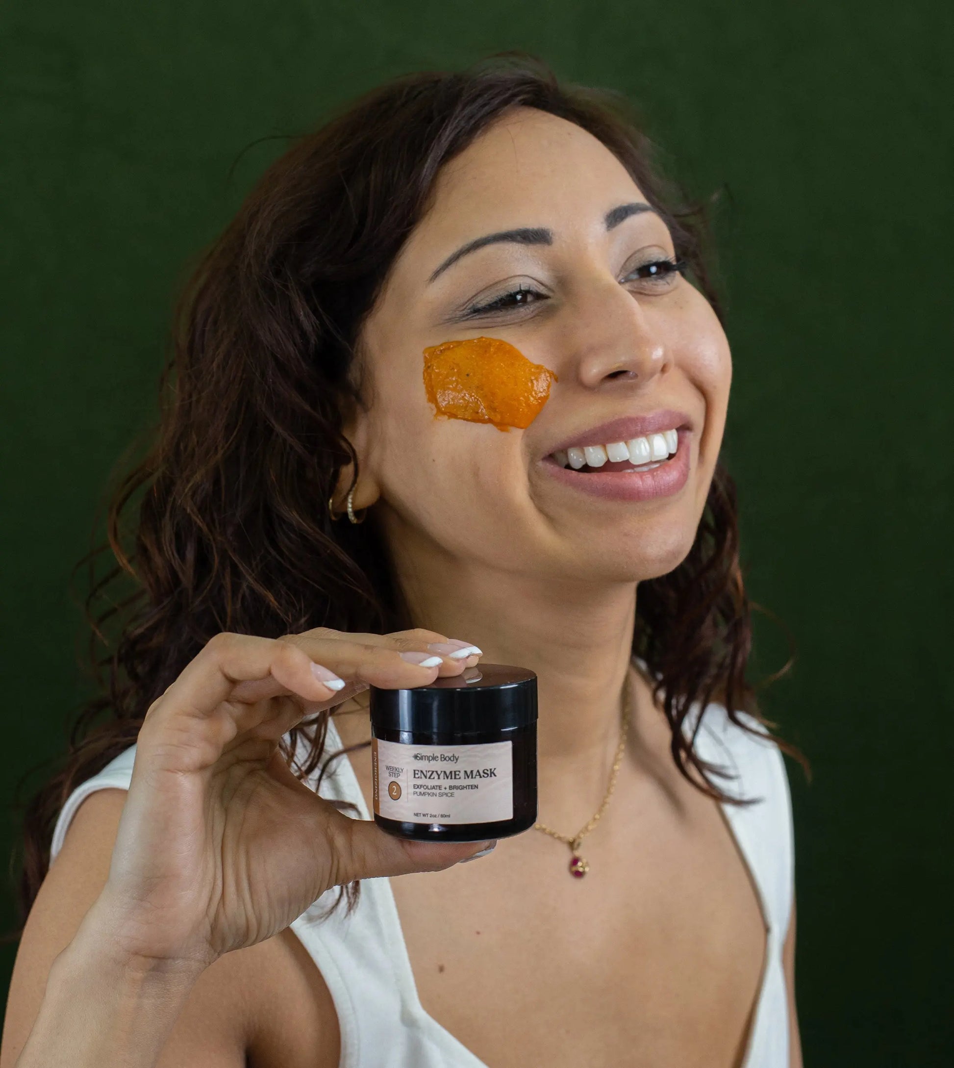 A woman looking off to the side with a swipe of Simple Body Pumpkin Enzyme Mask on her cheek which is a bright orange color holding a jar of the enzyme mask in one hand smiling with a dark green background.