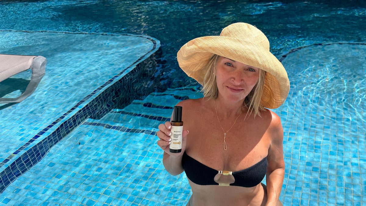 A woman in a black bikini top and a large straw sun hat stands in a tiled swimming pool, holding up a small amber bottle of Simple Body Brighten and Boost Skin Tonic. 