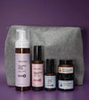 A Starter Skincare Kit for mature skin with a foaming face wash, Rosewater Toner, Nightly Face Serum, Beauty Balm, Face Cream, and a gray cosmetic bag on a purple background.