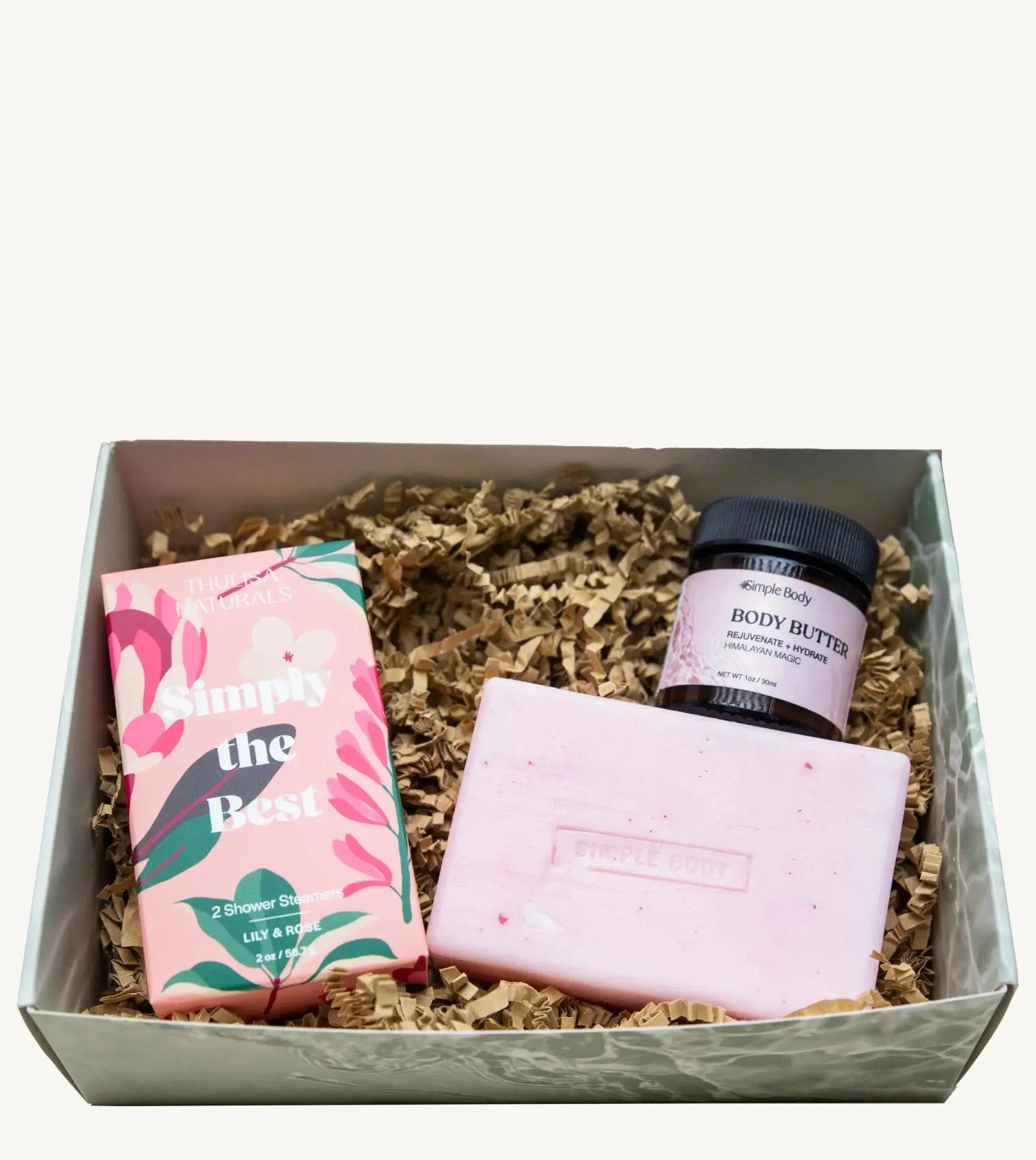 A gift box featuring a two-pack of shower steamers, handmade pink soap, and small jar of Simple Body Body Butter in Himalayan Magic with kraft colored shred on a white background.