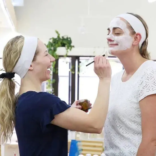 Two women applying Simple Body Kaolin Clay face masks to each other in a bright room.