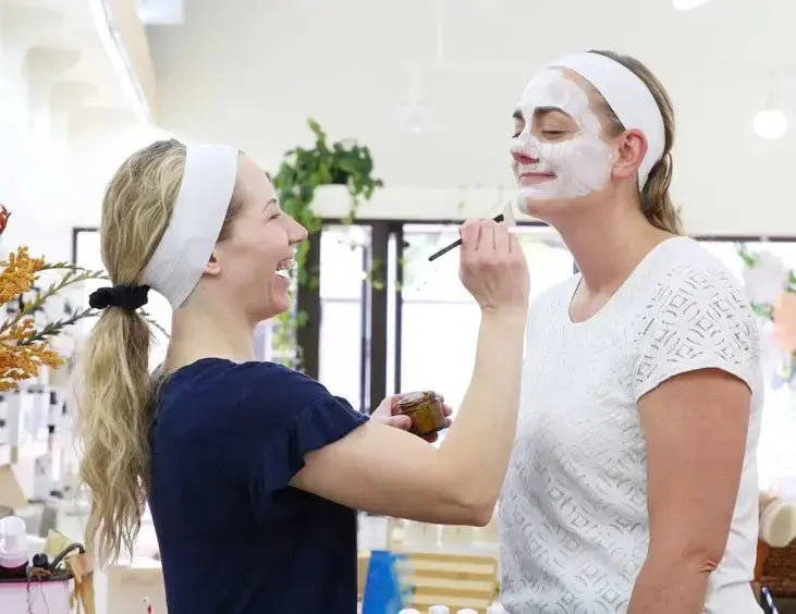 Two women applying Simple Body Kaolin Clay face masks to each other in a bright room.
