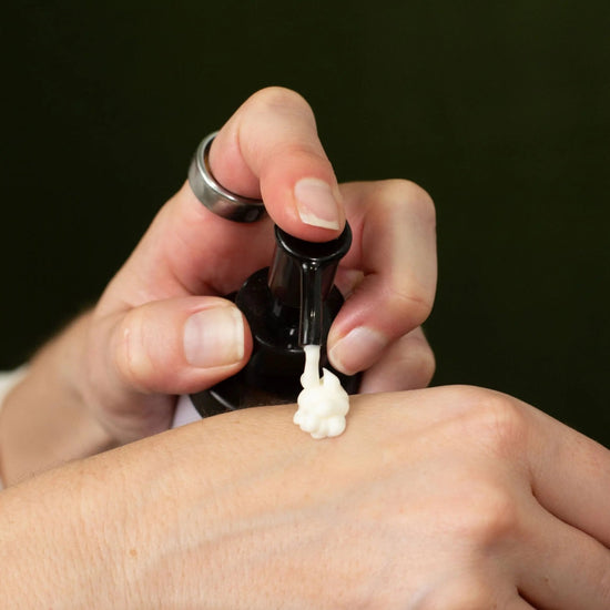 A hand holding a bottle of Simple Body Lotion pumping it onto a hand showing the creamy off white texture.
