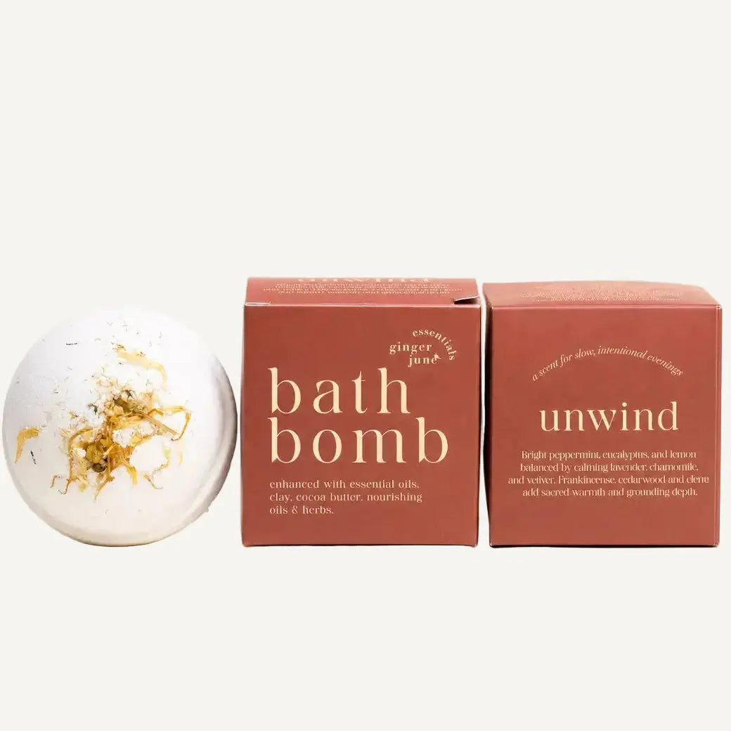 A white bath bomb by Ginger June Bath Co with herbs and botanicals sits beside two terracotta-colored product boxes on a warm cream background. The front box reads 'bath bomb' and 'ginger lime,' while the rear box reads 'unwind.