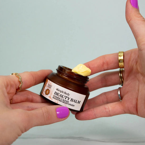 Person holding a jar of Simple Body Beauty Balm with a focus on the product--a Vitamin C yellow thick cream with brightly colored purple nails on a green background