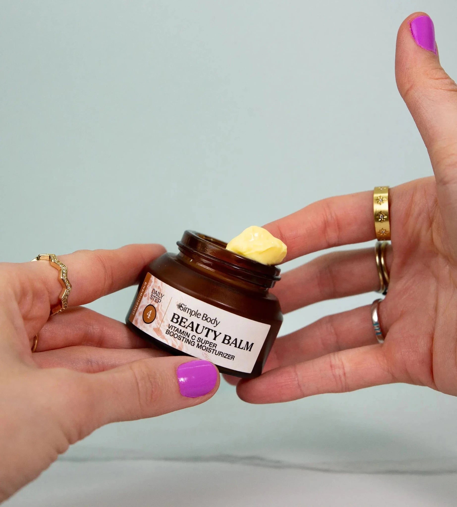 Person holding a jar of Simple Body Beauty Balm with a focus on the product--a Vitamin C yellow thick cream with brightly colored purple nails on a green background