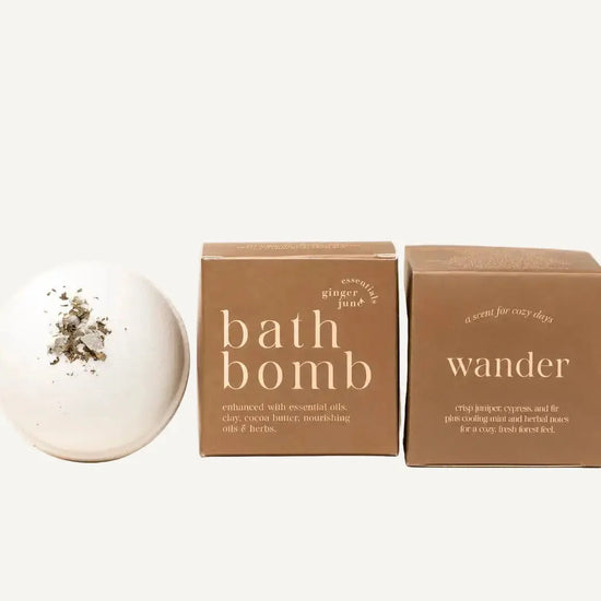 A white bath bomb from Ginger June Bath Co in Wander with dried botanicals on top sits next to two brown cardboard boxes labeled 'bath bomb' and 'wander' on a warm beige background.
