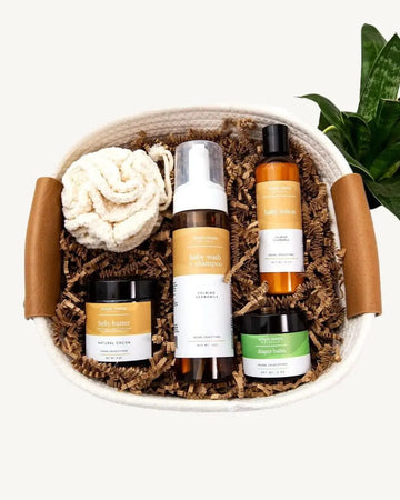 A Welcome Baby Gift Basket containing a Baby Wash, Baby Lotion, Belly Butter, and Diaper Balm, plus a cotton pouf, all beautifully arranged in a natural rope basket.