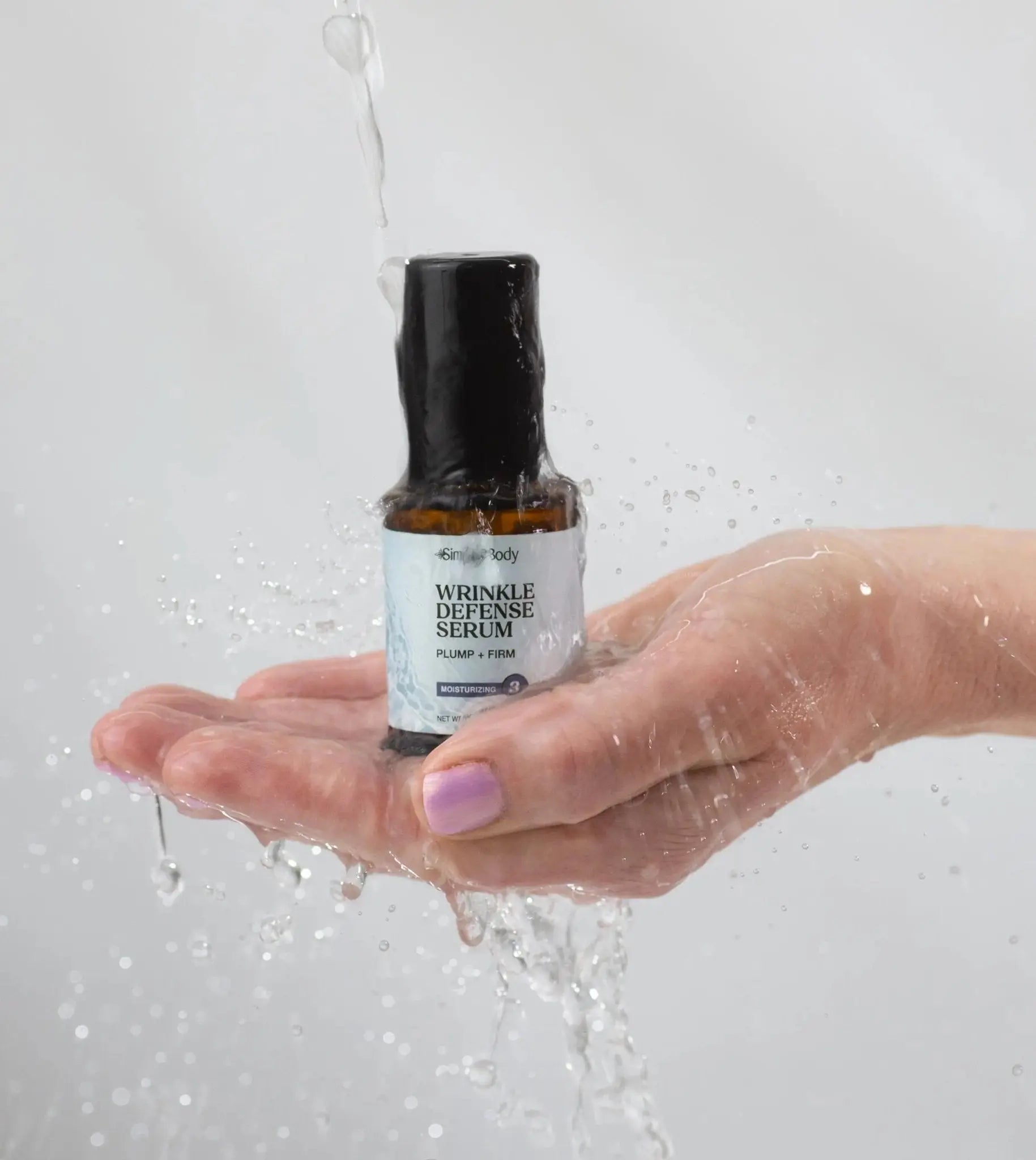 A woman's hand holding a Simple Body Wrinkle Defense Serum with water splashing down on it with a neutral background.